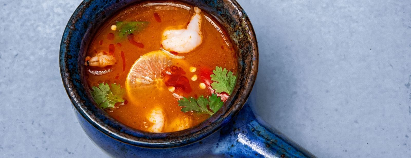 A bowl of soup with vegetables and herbs, served in a dark blue ladle-like bowl on a light textured surface at The Hamuse Hotel, Kodaikanal.