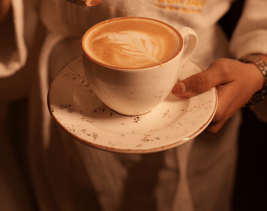 A person in a white robe holds a latte with foam art on a saucer, lifting a spoon. Warm, cozy ambiance with a focus on relaxation and indulgence.