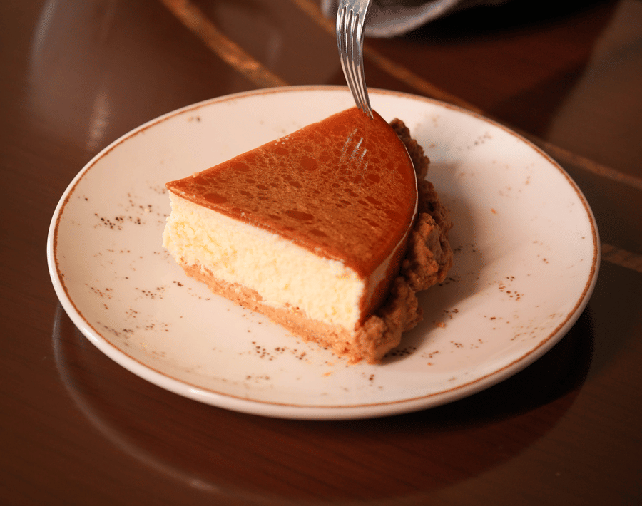 A person holds a fork over a caramel-topped cheesecake slice on a white plate. A cup of cocoa is nearby on the wooden table, creating a cozy feel.