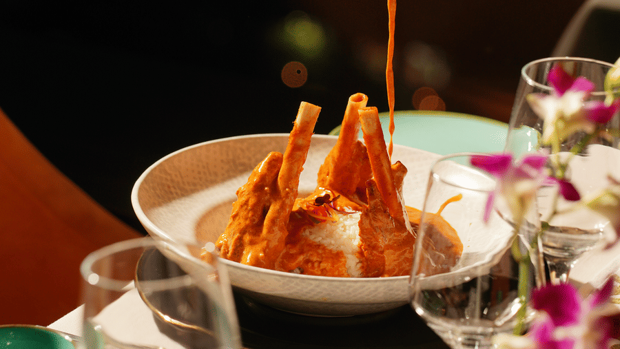A hand pours rich, orange curry sauce over a bowl with crispy pastry sticks. It's set on an elegant dining table with orchids and glassware, evoking warmth.