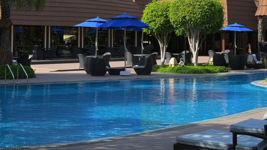 Outdoor swimming pool surrounded by lounge chairs and umbrellas, with a resort-style building and palm trees in the background, set against a mountainous landscape - Ananta Spa and Resort, Pushkar.