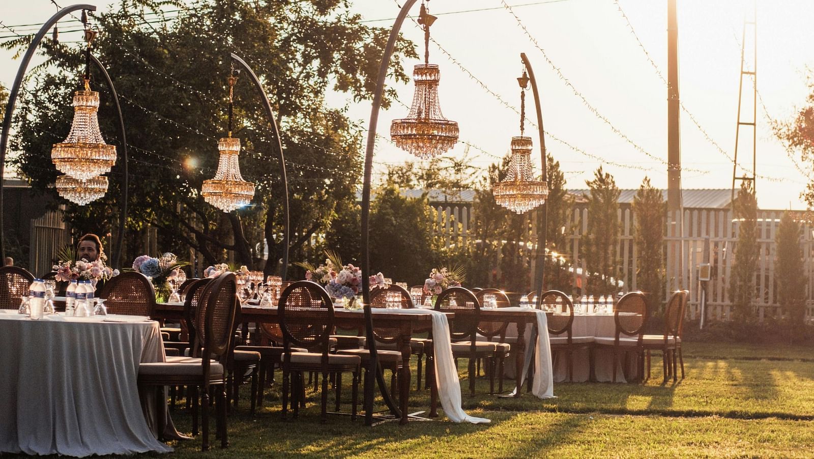 An outdoor lawn decorated with string lights, tableware and hanging lanterns at sunset.