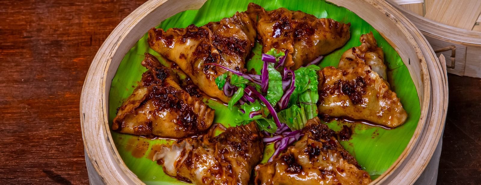 A bamboo steamer basket filled with appetizer garnished with green onions, surrounded by garlic bulbs and fresh scallions on a wooden table at The Hamuse Hotel, Kodaikanal.