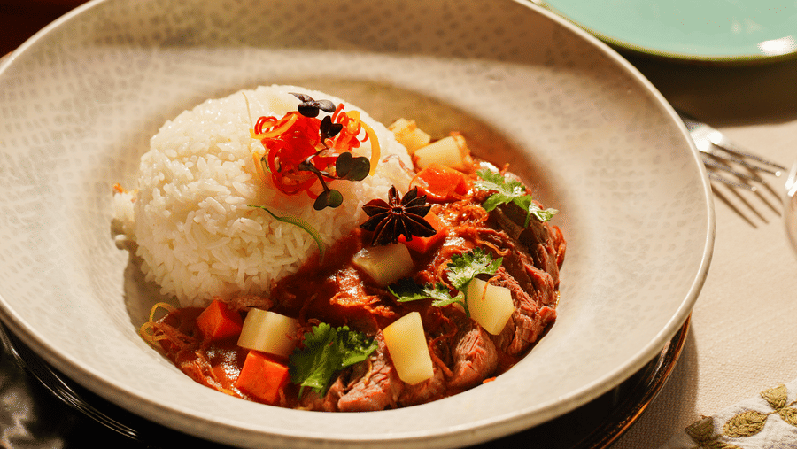 A plate of white rice topped with sliced red chili and herbs, alongside beef stew with vegetables and star anise, set on a patterned tablecloth.