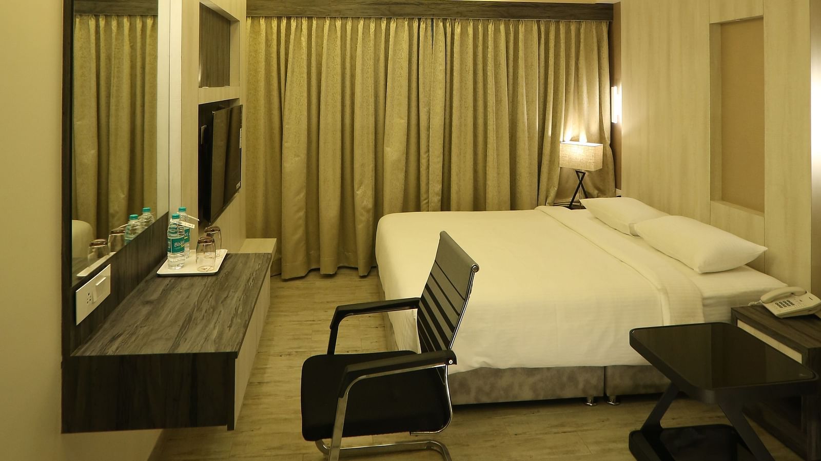 Executive Room featuring a bed with a dark headboard, a desk, and a chair, illuminated by warm lighting and complemented by dark curtains and a beige wall.