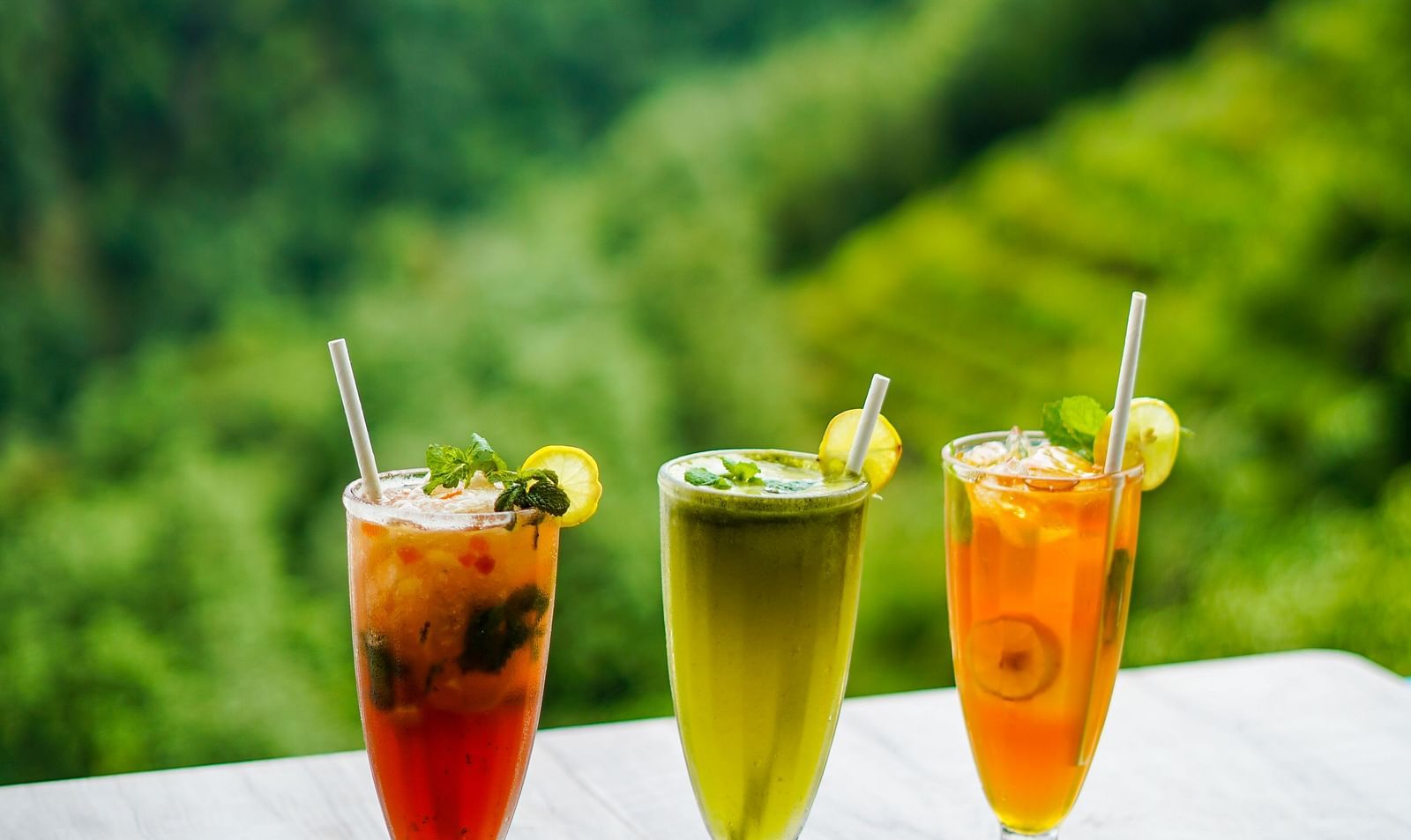 Three colourful beverages in glasses on a white table with a lush green mountain in the background | Sukama Resort & Spa, Nainital