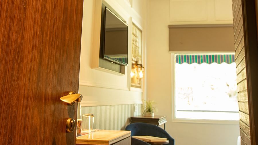 A hotel room door half-open to a cozy room with dark wood accents, a bed, seating area, and bright windows.