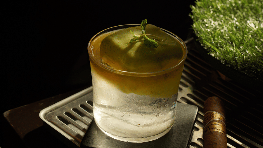 A person carefully cuts a cigar above a cocktail garnished with a fresh lime slice, surrounded by dark foliage, creating a sophisticated, moody ambiance.