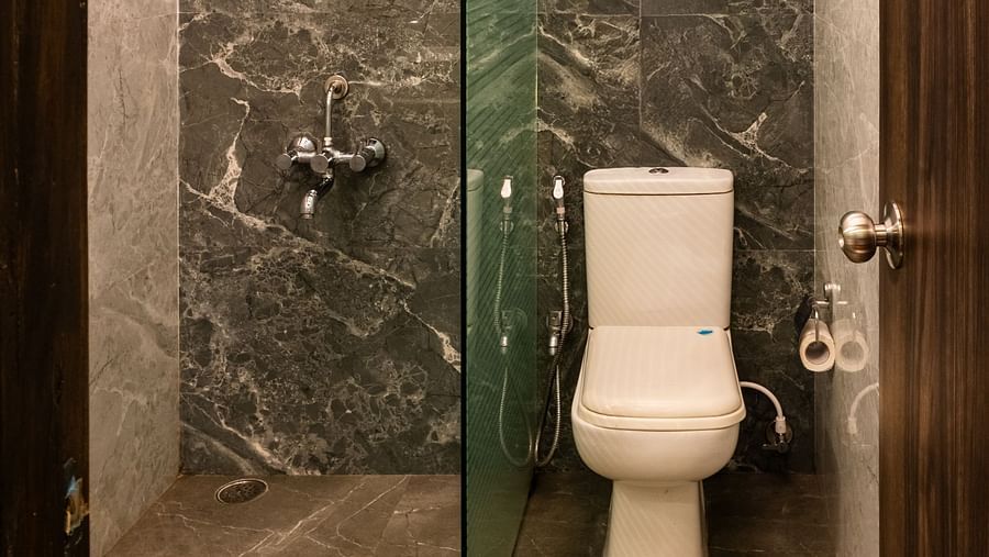 Compact and stylish bathroom with marble-textured walls and flooring, a glass partition for the shower area, and a Western toilet under a designer ceiling - The Grand Legacy Resort & Spa