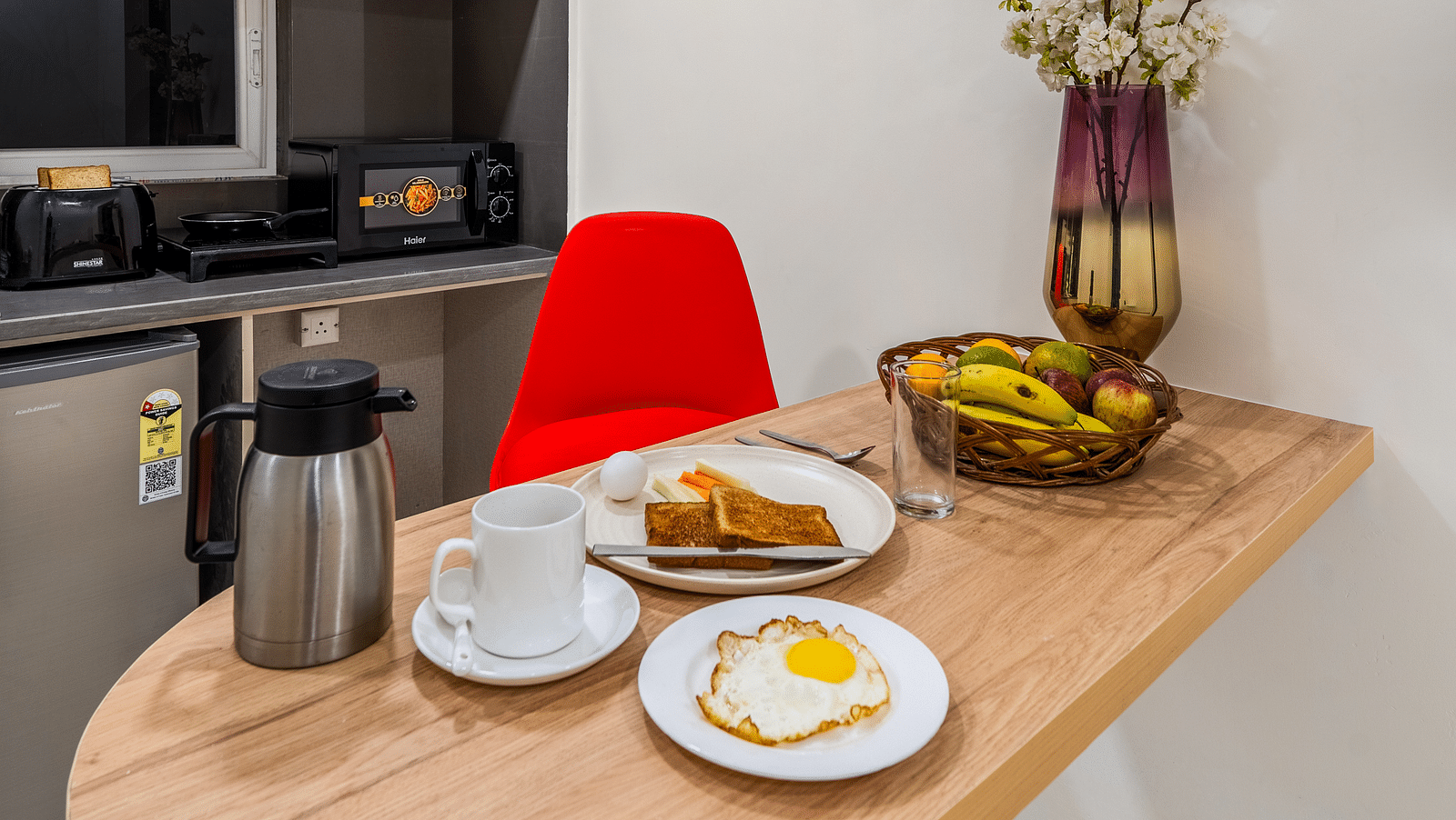 Image of a wooden dining table that is fixed to the wall and has a flower vase, white plates with food and a kettle | Eminent Suites & Apartments, Hi-Tech City