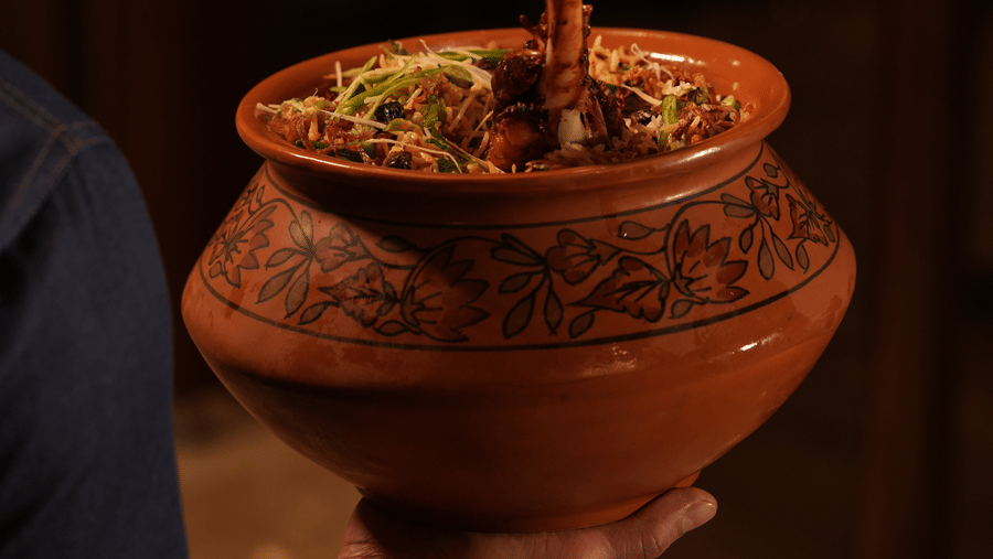 A person holds a decorated clay pot of biryani topped with a protruding meat bone. The setting is warm and inviting, emphasizing culinary tradition.