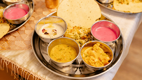 A traditional thali meal of various curries, pink yogurt, and flatbread served on steel platters for two people seated at a table.