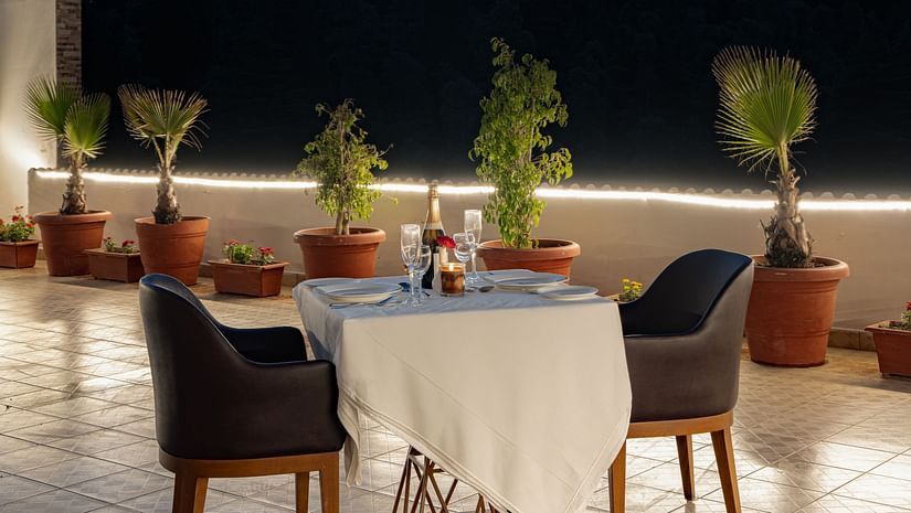 An outdoor dining setup at Clarks Inn, Near Kainchi Dham at dusk features a set table for two, surrounded by potted plants and soft string lighting.