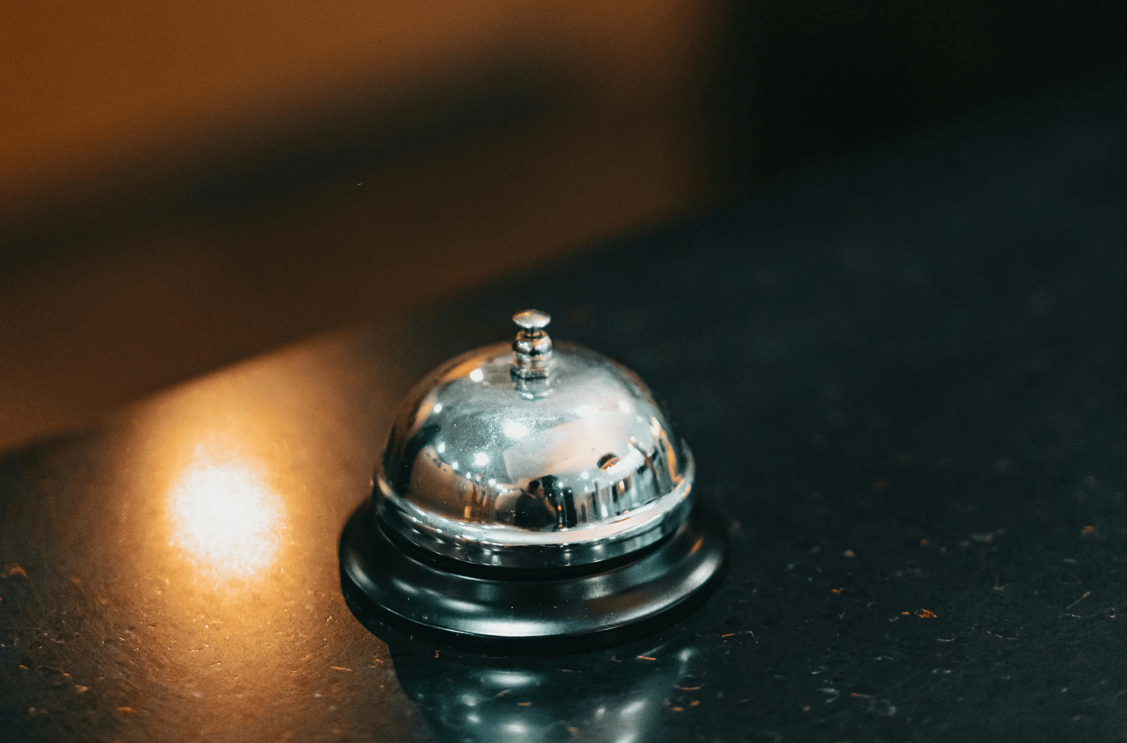 A close up of a shiny, silver service bell on a dark, reflective surface, illuminated by a warm light in a dimly lit room.