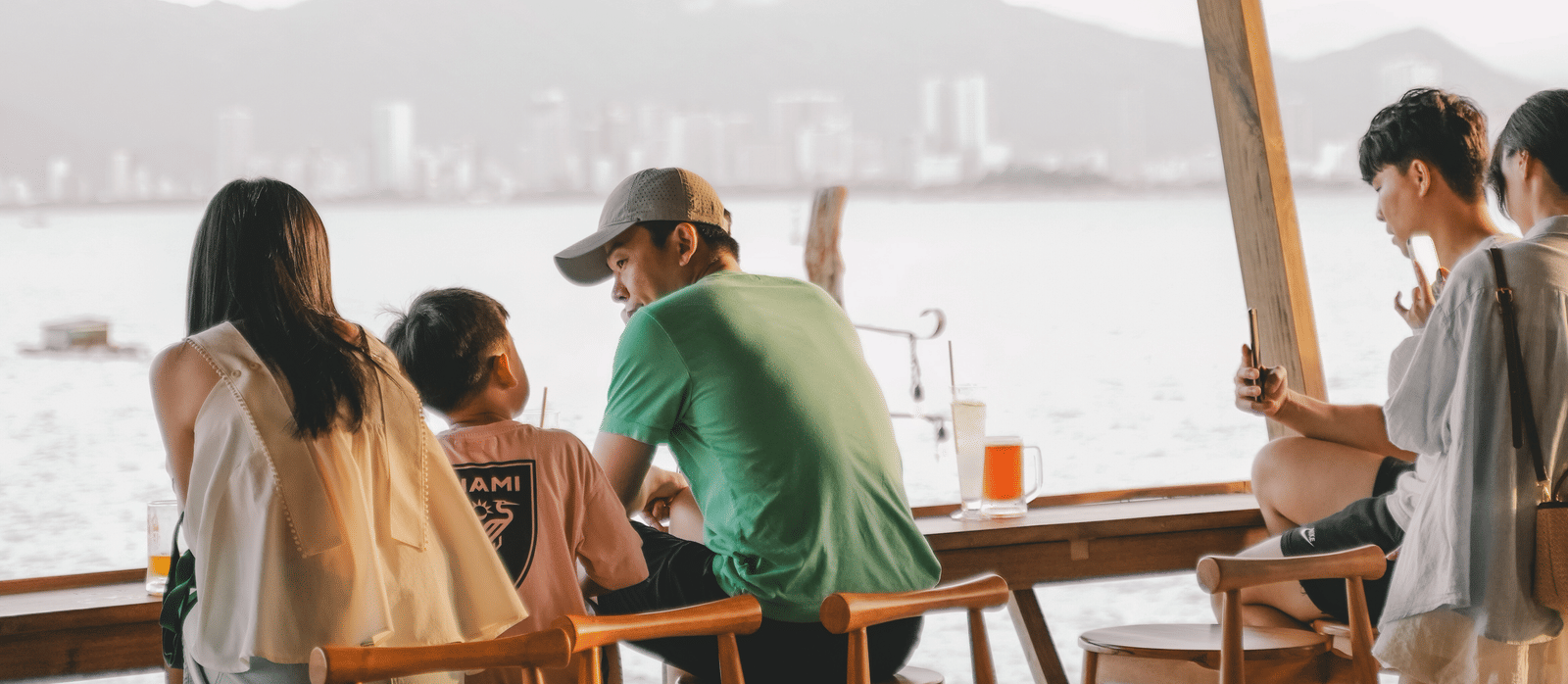 People sitting a wooden furnished restaurant which has ceiling lights and a river view.