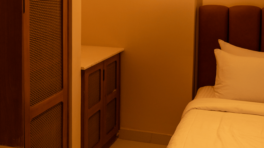 A single bed with white bedding is seen next to a tall wooden wardrobe and a modern vertical hanging light at Avanya Resorts N Retreat.