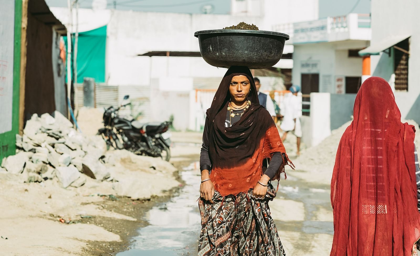 Two women walk down a street, one carrying a bowl on her head, in a village setting with buildings and a bright sky- Shahpura Bagh.