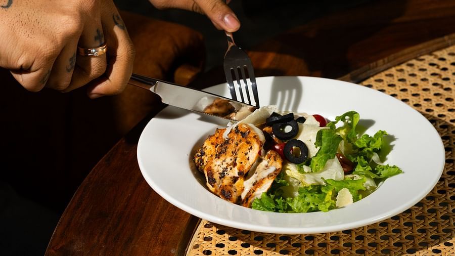 A person's hands holding utensils over a plate of food, set on a table with a woven placemat at The Qualia Resort Club & Brewery, Udaipur.
