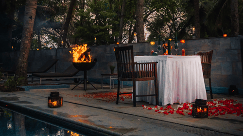 A romantic, private dinner set up poolside at dusk, surrounded by lush tropical greenery and tall palm trees.