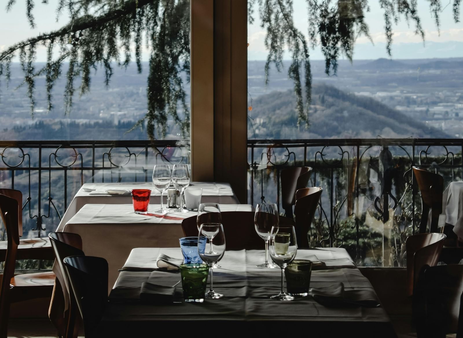 Indoor restaurant dining area with set tables, glassware, and a scenic view of mountains and trees visible through large windows in the background.