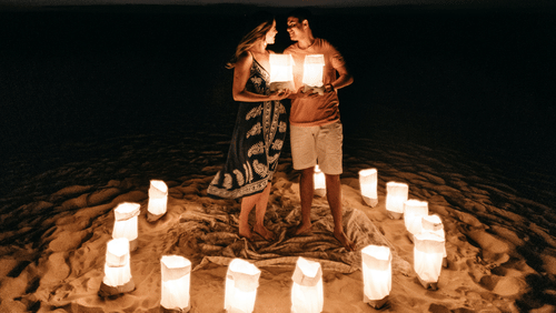 Two people standing on a beach in a circle of white lanterns.