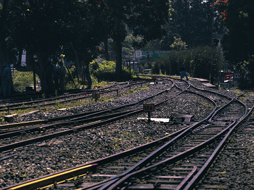 Railway tracks curve through lush greenery beneath a bright sky in the Nilgiri hills.