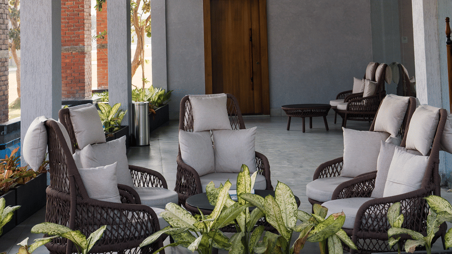 An open-sided covered area featuring pillars, containing multiple dark woven armchairs with white seat cushions arranged around small tables at Hotel Sonar Bangla, Ajodhya Hills.