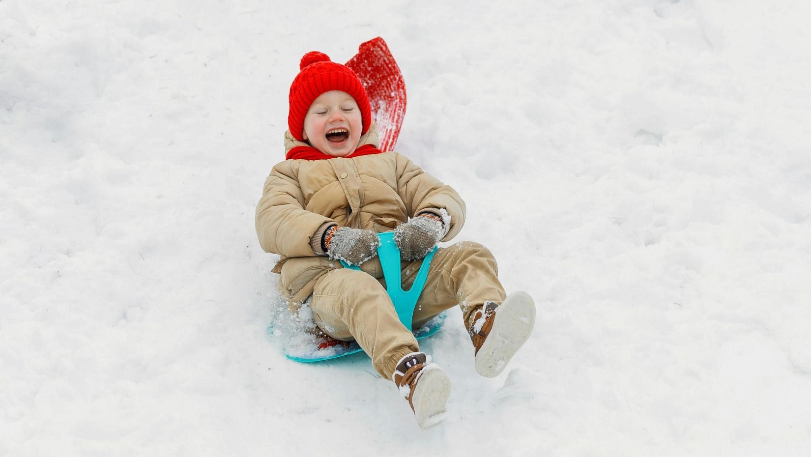 Two children play in a snowy landscape, one sledding down a hill, the other holding a sled in the background.