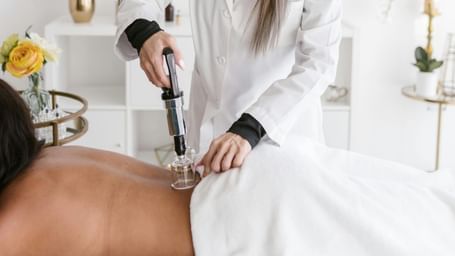  A person in a white lab coat performs cupping therapy on a person lying on their stomach on a massage table. A cupping gun is being used.