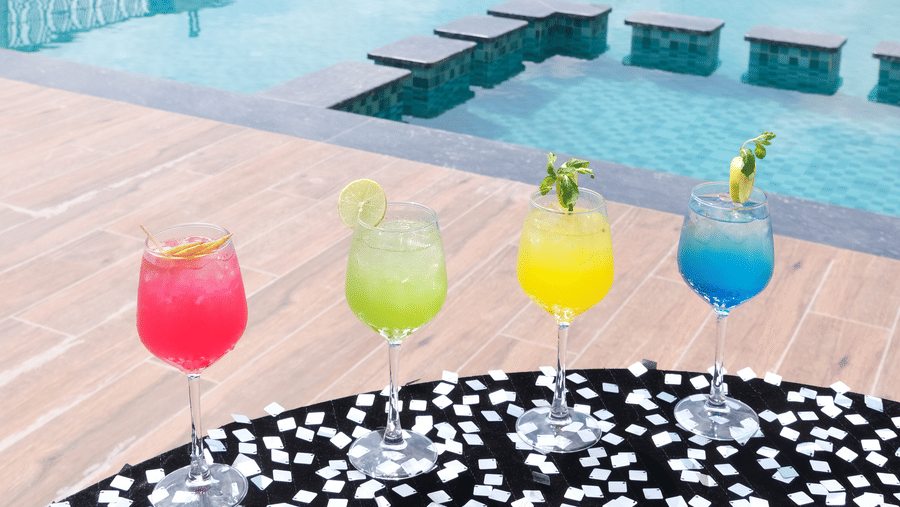 an outdoor swimming pool with a row of drinks on a patterned table in the foreground | Pride Premier Alwar
