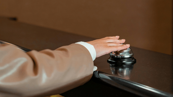 Person pressing a silver service bell on a wooden front desk counter.