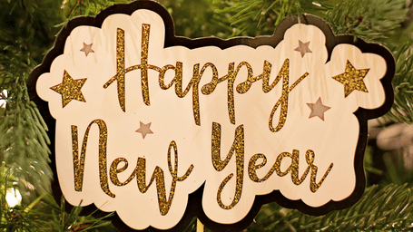 Happy New Year sign and a clock ornament, showing midnight, hanging on a blurred Christmas tree.