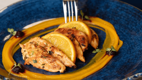 Blue plate with grilled chicken breast slices, orange slices, pumpkin puree swirl, microgreens, and berry garnish with a fork at V FIVE HOTEL.