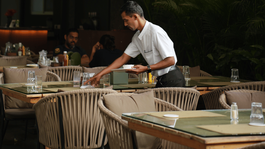 A restaurant area at Oceanature Resort, Alibaug with a server arranging tables beneath hanging woven lamps.