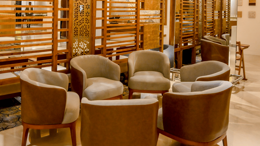 A lounge area features 5 grey armchairs arranged in a circle next to a patterned partition wall under hanging pendant lights at Tiara by MPS.