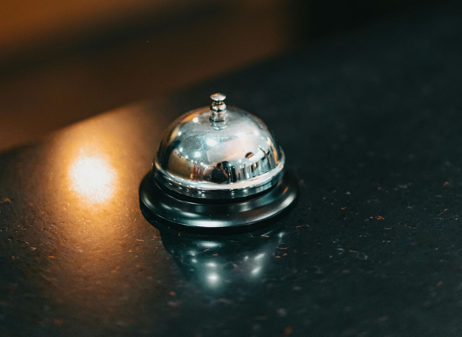 A close-up of a shiny, silver service bell on a dark, reflective surface, illuminated by a warm light in a dimly lit room.