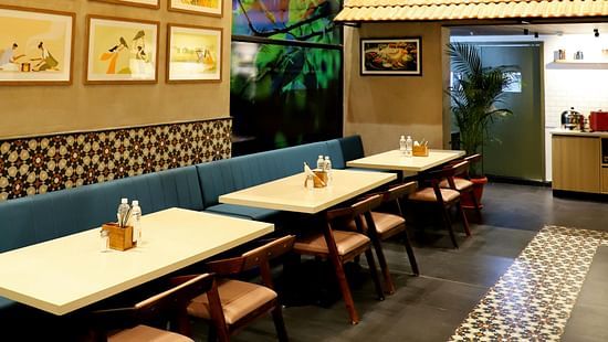 The interior of a casual restaurant or cafe with wooden tables, booth seating, hanging light fixtures, and framed pictures on the walls.