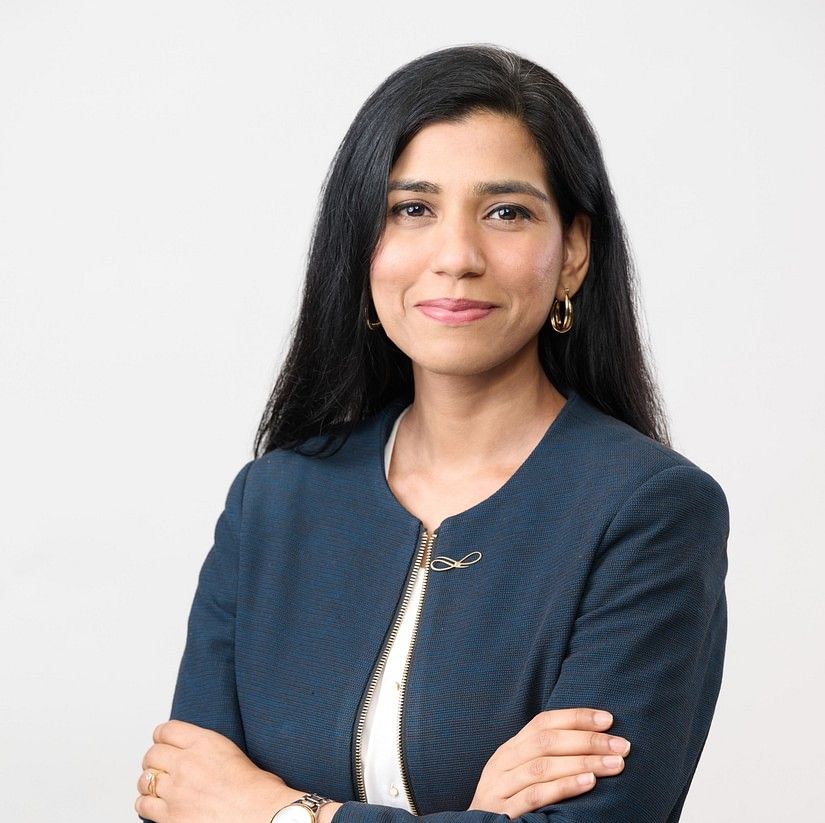 Manisha with long black hair, wearing a navy blazer and white shirt, standing with arms crossed, smiling slightly.