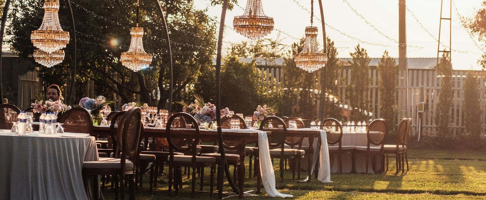 An outdoor lawn decorated with string lights, tableware and hanging lanterns at sunset.