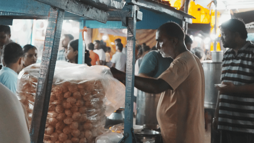 A phuchka stand with the vendor making them to give it to the people with a lit building in the background