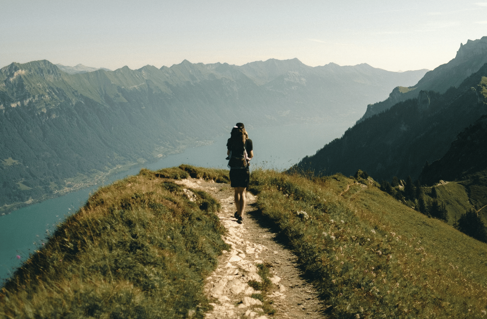 A lone hiker standing on a grassy mountain ridge looking down at a vast blue lake nestled in a valley.