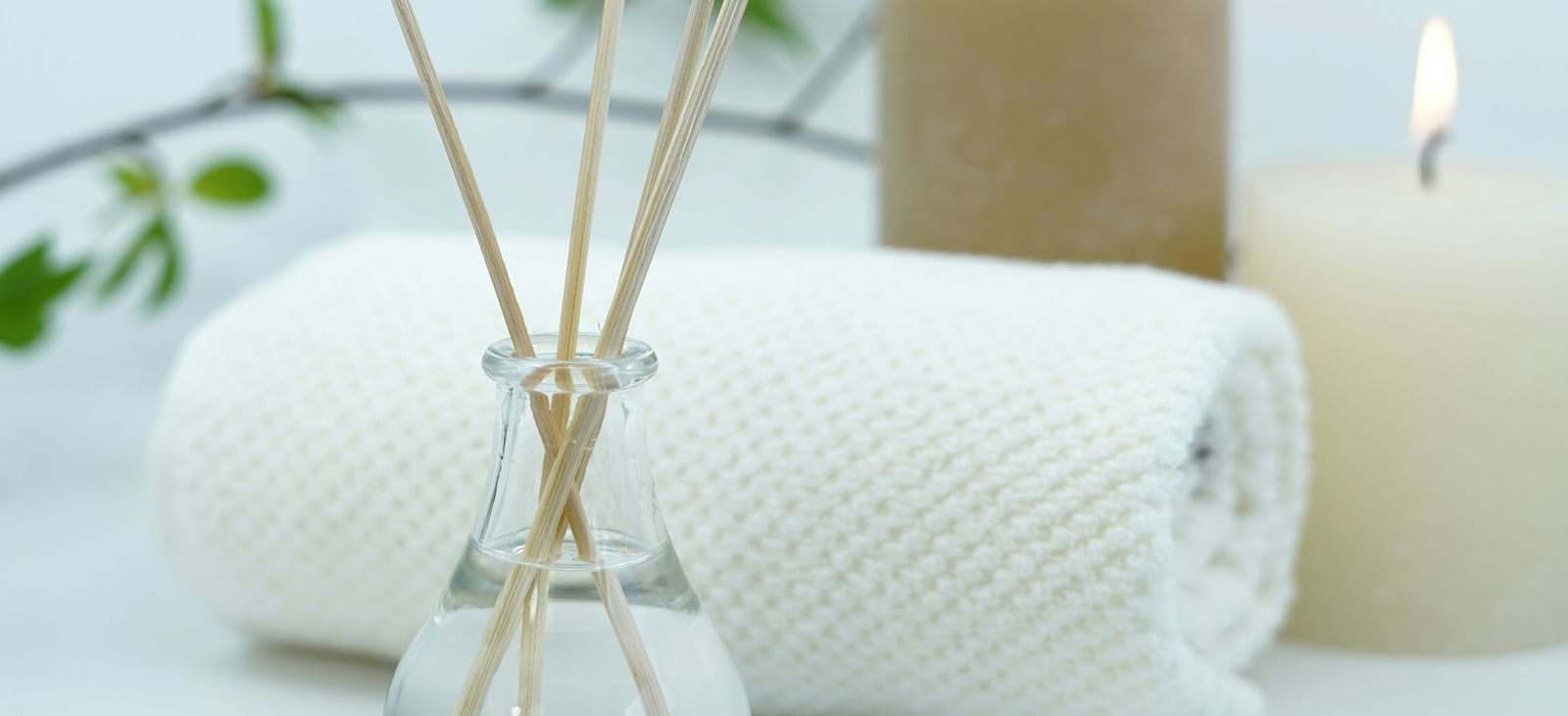 An arrangement of spa or wellness items, including a reed diffuser, a lit candle, and rolled white towels, set against a light background.