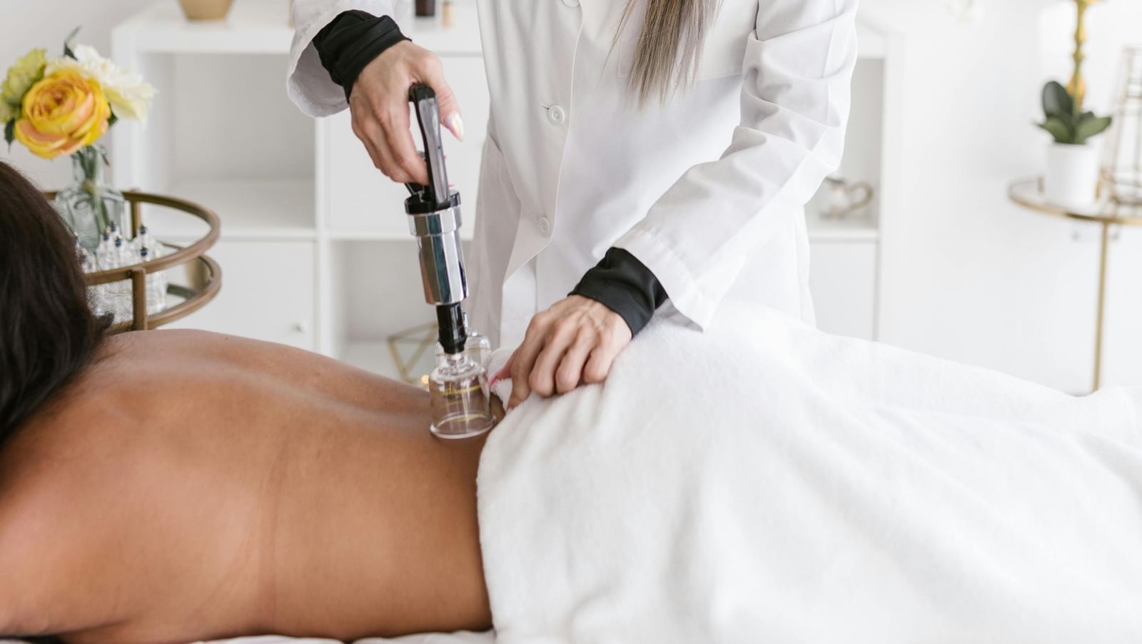  A person in a white lab coat performs cupping therapy on a person lying on their stomach on a massage table. A cupping gun is being used.