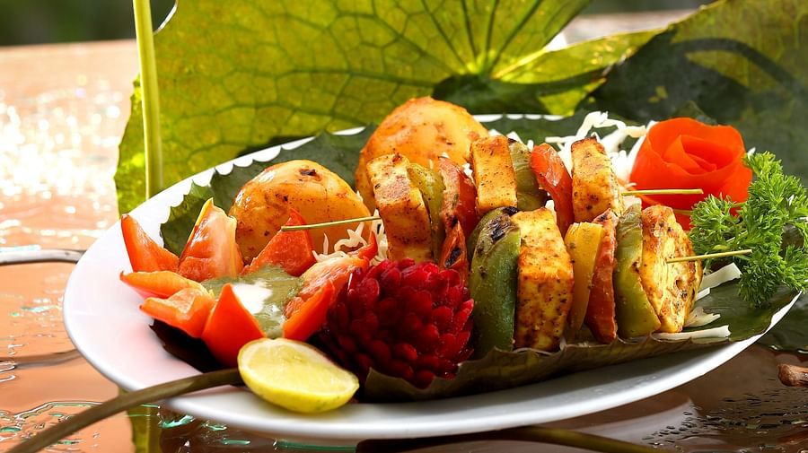 A vegetarian platter sits next to a lotus leaf and bud, while a seafood dish is displayed on the table, with 'Aambal' illuminated on the wall in the background at Uday Backwater Resort