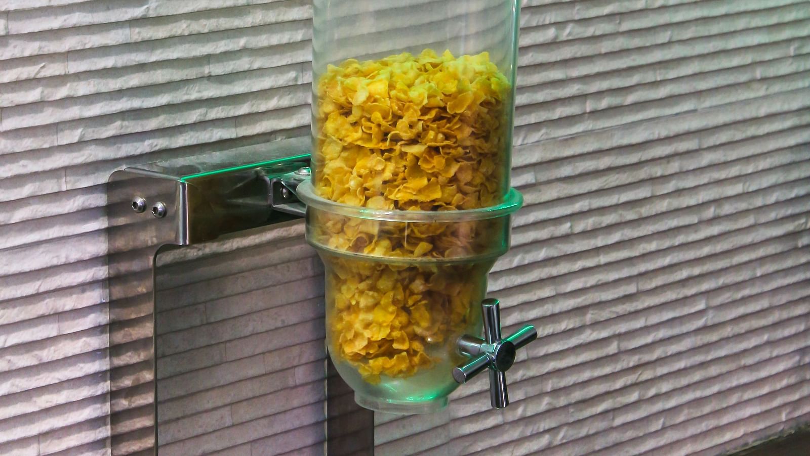 A clear plastic dispenser filled with cereal, mounted on a textured wall with green lighting above. - Hotel Diamond Plaza, Chandigarh.
