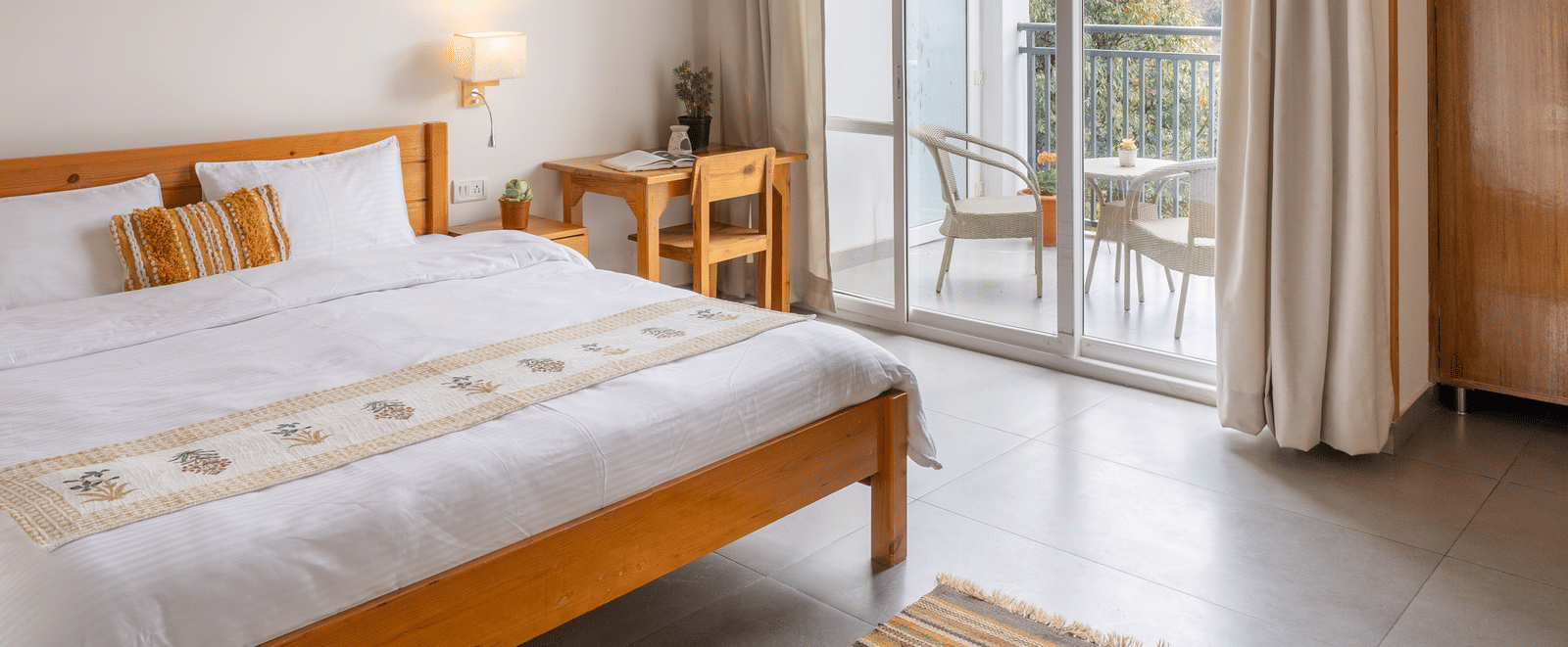 A cropped image of a Ziran Retreat bedroom with a bed, bedside table, and a large window with white curtains, showing a partial view of the room and the window.