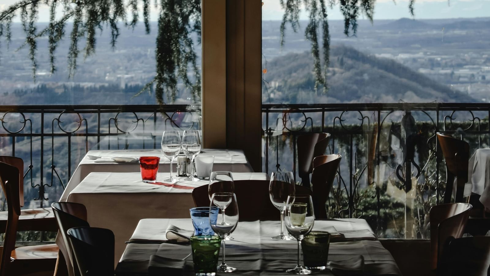Indoor restaurant dining area with set tables, glassware, and a scenic view of mountains and trees visible through large windows in the background.