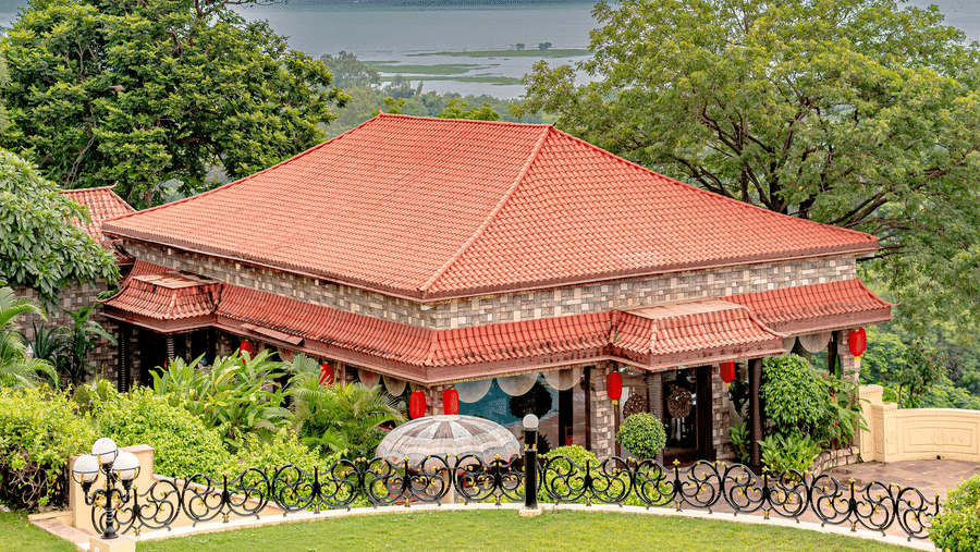 An exterior view of the Ming Dynasty restaurant at Noor-Us-Sabah Palace, Bhopal, showing the traditional red-roofed structure set amidst lush gardens under a cloudy sky.