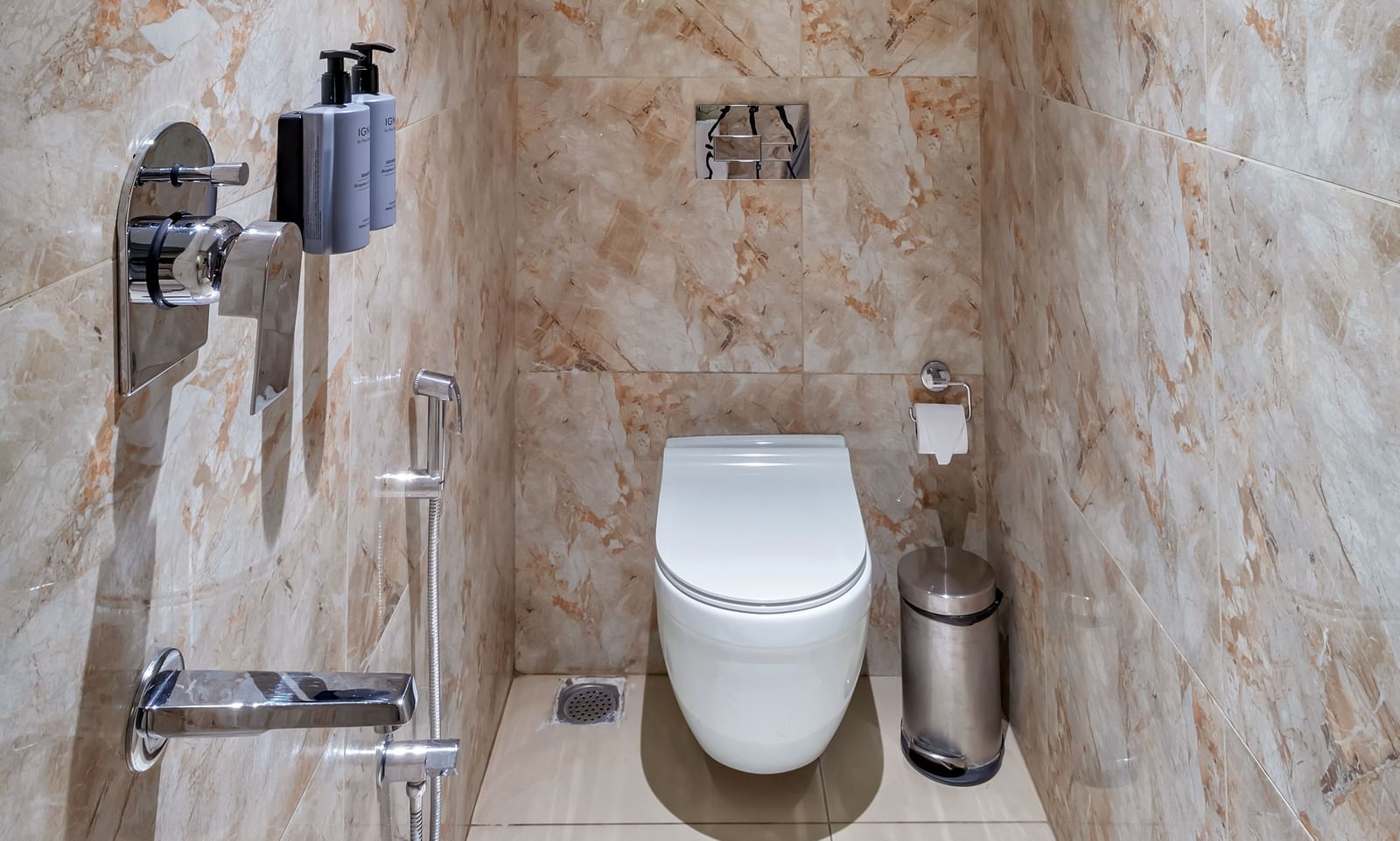 A narrow bathroom with marble-patterned walls, a white toilet, a metal trash can, and chrome fixtures | The Citi Residenci Hotel Banskopa