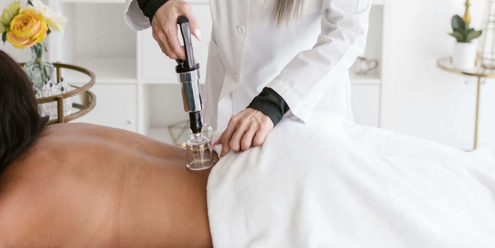  A person in a white lab coat performs cupping therapy on a person lying on their stomach on a massage table. A cupping gun is being used.